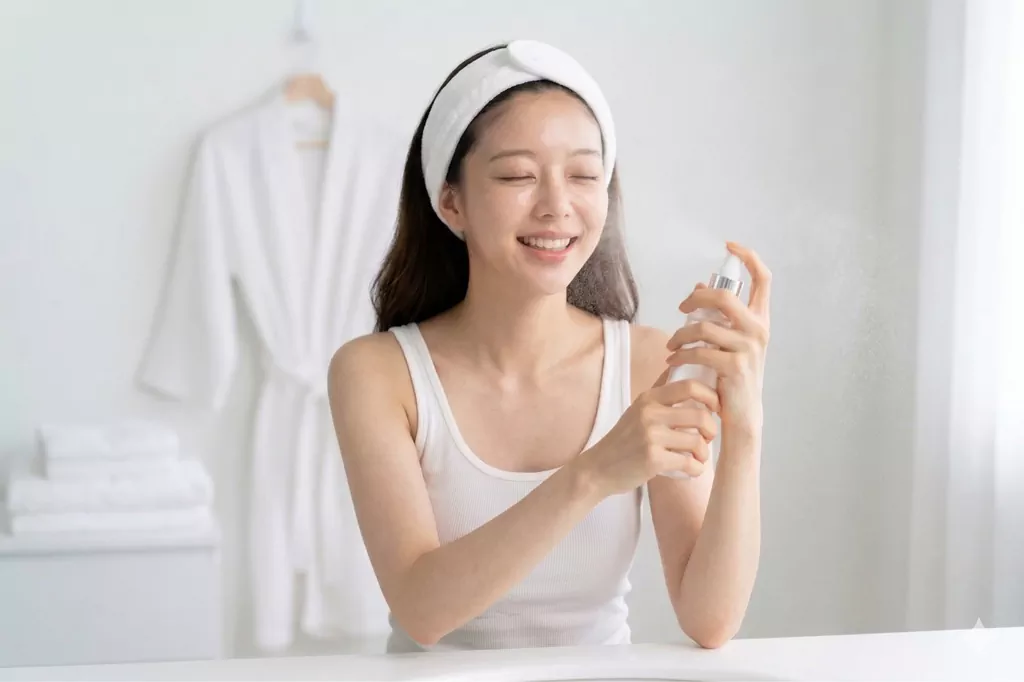 5 Steps to a Flawless Skin Flooding Routine for That Iconic Glass Skin Glow 1 A hand spraying a hydrating facial mist to create damp skin, which is the vital first step in a successful skin flooding routine.