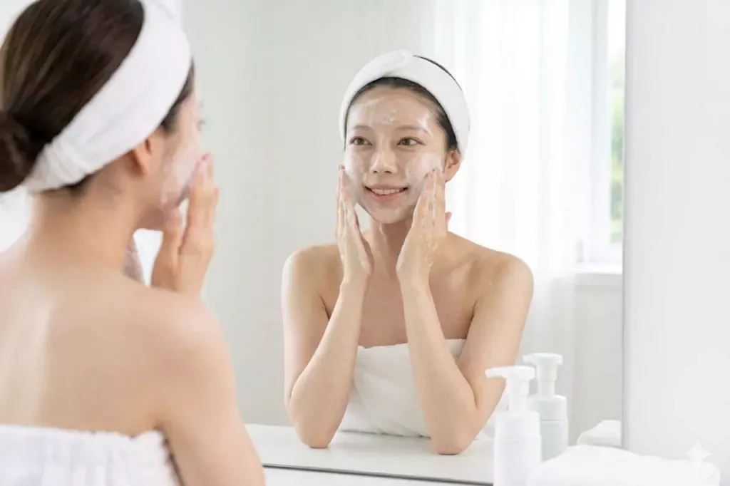 K-Beauty Double Cleansing Guide: The Ultimate Reset for Glowing Skin 2 The second step of the **K-Beauty Double Cleansing Guide** showing a woman washing her face with a gentle white foam cleanser in front of a mirror.